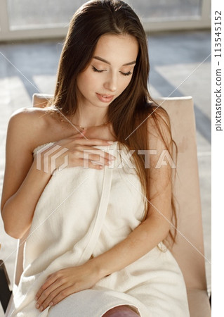 Young woman in cosmetology cabinet. Brunette woman sitting in a bright room with big windows. Girl wraps her body in white towel. 113545152
