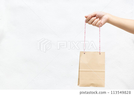 Hand of business woman hold brown paper shopping bag on white concrete background Hand of business woman hold brown paper shopping bag on white concrete background 113549828