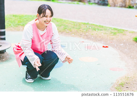 A nursery teacher playing with a child in front of the playground equipment in the park 113551676