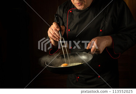 A cook in uniform holds a frying pan with a hot fried egg and a fork. Low key concept of cooking scrambled eggs. Place for advertising on a black background 113552025