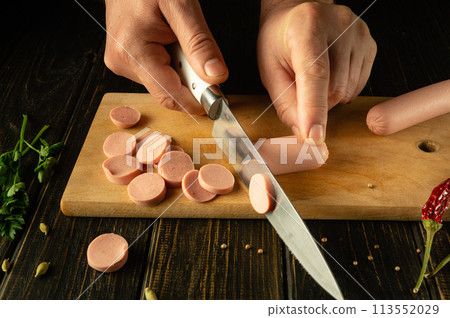 The cook cuts vienna sausage on a wooden cutting board. Preparing delicious sandwiches for breakfast on the kitchen table in a restaurant 113552029