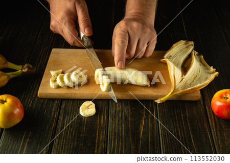 The cook cutting banana on a wooden boards. Low key concept of preparing a fruity breakfast dish on the kitchen table. Advertising space 113552030