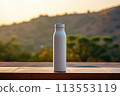 A plastic bottle filled with liquid is placed on a wooden table against the backdrop of a majestic mountain and clear blue sky, surrounded by lush green grass 113553119