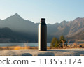 A black water bottle is placed on a rock overlooking a stunning natural landscape with mountains in the background, near a serene lake reflecting the blue sky 113553124