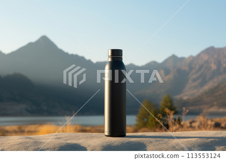 A black water bottle is placed on a rock overlooking a stunning natural landscape with mountains in the background, near a serene lake reflecting the blue sky 113553124