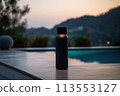A black bottle rests on a wooden table overlooking a serene swimming pool, surrounded by lush plants with mountains in the horizon under a clear sky 113553127