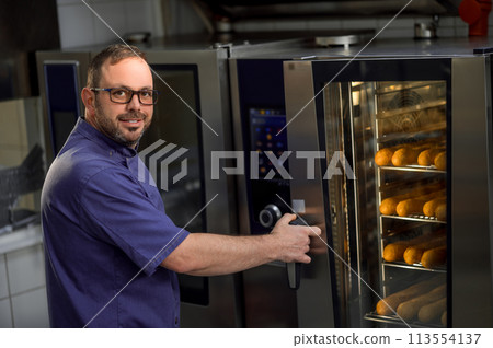 Baker in kitchen near oven with baguettes in bakehouse 113554137