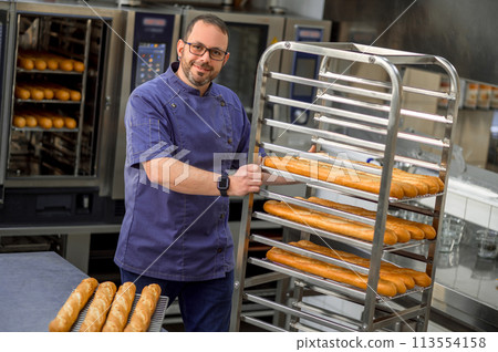 Man baker in uniform baking baguettes with bread shelves on background Man baker in uniform baking baguettes with bread shelves on background 113554158