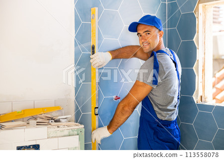 Senior master in blue construction overalls accepts correct laying of tiles on wall using level Senior master in blue construction overalls accepts correct laying of tiles on wall using level 113558078