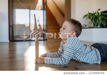 Boy blowing into blades of wind turbine model. Concpet of renewable energy sustainable lifestyle for next generations. Boy blowing into blades of wind turbine model. Concpet of renewable energy sustainable lifestyle for next generations. 113564354