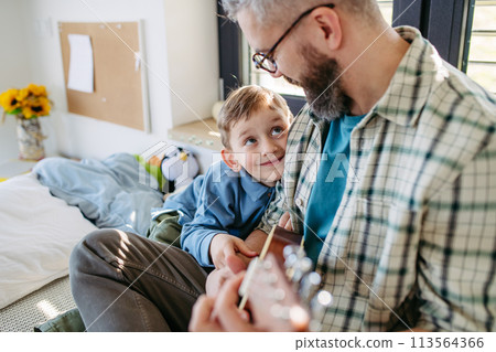 Father teaching boy to play on acoustic guitar. Mesmerized son listening dad making music. Concept of Fathers Day and fatherly love. 113564366