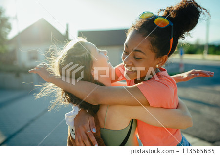 Teenager girl best friends greeting each other, hugging. Girls spending time outdoors in city during warm summer holiday day. 113565635
