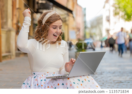 Caucasian happy woman working on laptop celebrate success win money sitting on urban street in city Caucasian happy woman working on laptop celebrate success win money sitting on urban street in city 113571834