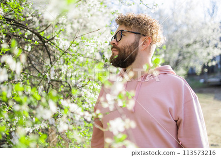 Man with beard and mustache on smiling face near sakura flowers or blooming spring tree. Soft and gentle concept. Bearded man with stylish haircut with flowers on background, close up. Hipster near 113573216