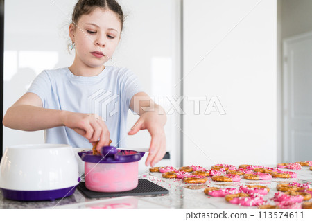 With focused attention, this budding culinary artist dips pretzels into a pot of melted chocolate, creating sweet delights in the warmth of a well-lit home kitchen. 113574611