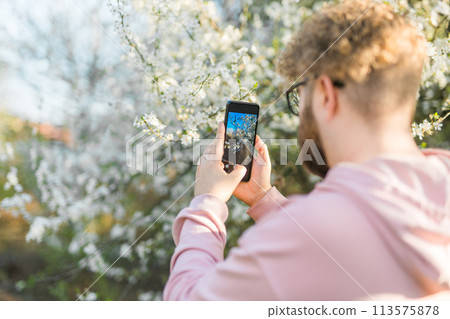 Man holding mobile phone and take photo blooming spring cherry and apples trees in sunlight. Smartphone photo for social media. Copy space Man holding mobile phone and take photo blooming spring cherry and apples trees in sunlight. Smartphone photo for social media. Copy space 113575878