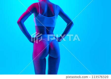 Rear view photo of toned female body, buttocks in neon pink light against gradient blue background. Model in lingerie put hands on hips. Rear view photo of toned female body, buttocks in neon pink light against gradient blue background. Model in lingerie put hands on hips. 113576320