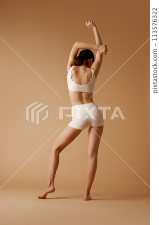 Rear view photo of young woman posing in sportswear that emphasizes her perfect curves against pastel beige background. Rear view photo of young woman posing in sportswear that emphasizes her perfect curves against pastel beige background. 113576322