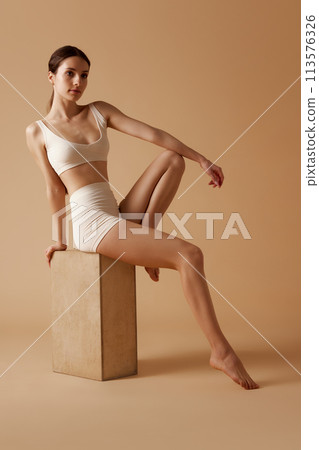 Young, slim female model posing sitting on wooden cube in white sportswear against pastel beige studio background. Young, slim female model posing sitting on wooden cube in white sportswear against pastel beige studio background. 113576326