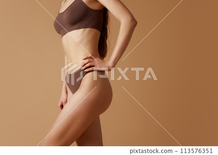 Cropped photo of slim, young woman posing in brown lingerie holding hands on hips against sandy background. 113576351