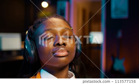College student sitting laid back after working on class project, relaxing with ambience music on headphones at home desk. African american girl chilling with modern tunes on headset. Camera B. College student sitting laid back after working on class project, relaxing with ambience music on headphones at home desk. African american girl chilling with modern tunes on headset. Camera B. 113577158