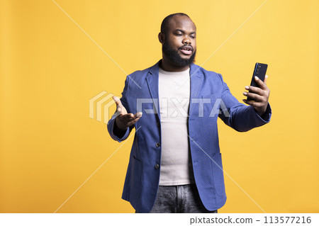 Annoyed man having unpleasant conversation with friends during teleconference meeting using smartphone, studio background. Hostile person having uncivil chat with mates during online videocall 113577216