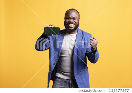 African american man excitedly celebrating while presenting isolated screen phone, studio background. Ecstatic BIPOC person holding copy space mockup cellphone used for advertising brands 113577277