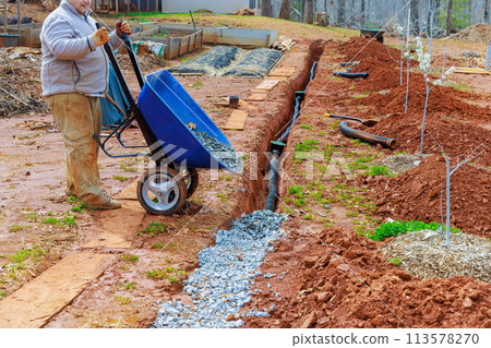 From wheelbarrow, worker fills overflowing drainage pipe with crushed stone From wheelbarrow, worker fills overflowing drainage pipe with crushed stone 113578270