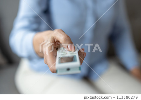 Close-up of a hand holding and pressing buttons on an air conditioner remote control, focusing on indoor temperature regulation. 113580279