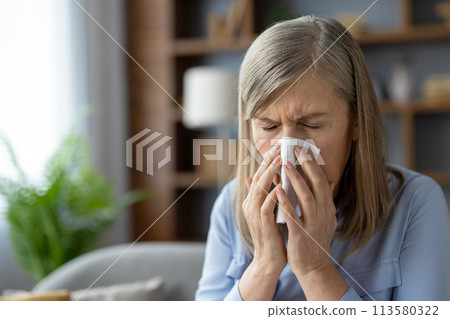 A mature woman experiencing cold symptoms, sneezing into a tissue while sitting in a cozy living room. Health and wellness concept. A mature woman experiencing cold symptoms, sneezing into a tissue while sitting in a cozy living room. Health and wellness concept. 113580322