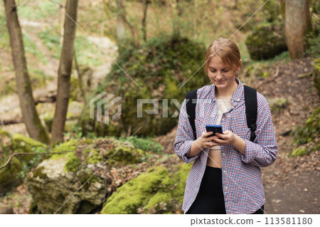 Girl tourist walking in forest and using smartphone. Young female with backpack hiking on the road in nature. Beautiful 30s woman walking on nature in sport clothes. High quality photo 113581180