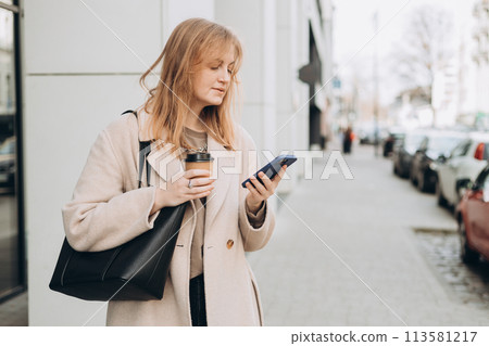 Beautiful smiling female with coffee cup using smartphone and standing outdoors. Phone Communication. Happy cheerful young woman walking on city street, Urban lifestyle concept. Traveler Beautiful smiling female with coffee cup using smartphone and standing outdoors. Phone Communication. Happy cheerful young woman walking on city street, Urban lifestyle concept. Traveler 113581217