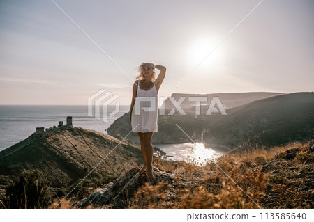 A blonde woman stands on a hill overlooking the ocean. She is wearing a white dress and she is enjoying the view. 113585640