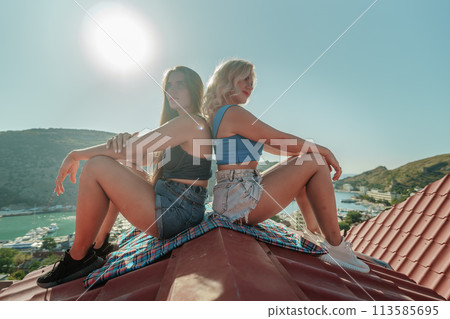 Two women sitting on a red roof, enjoying the view of the town and the sea. Rooftop vantage point. In the background, there are several boats visible on the water, adding to the picturesque scene. 113585695