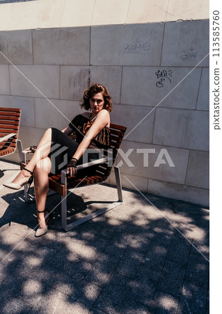 Portrait of a woman on the street. An attractive woman in a black dress is sitting on a bench outside. 113585760