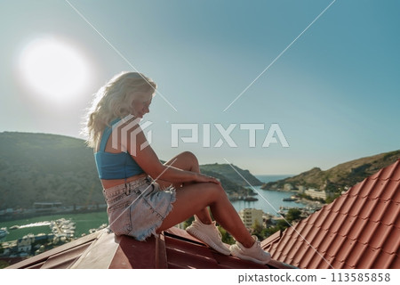Woman sits on rooftop, enjoys town view and sea mountains. Peaceful rooftop relaxation. Below her, there is a town with several boats visible in the water. Rooftop vantage point. Woman sits on rooftop, enjoys town view and sea mountains. Peaceful rooftop relaxation. Below her, there is a town with several boats visible in the water. Rooftop vantage point. 113585858