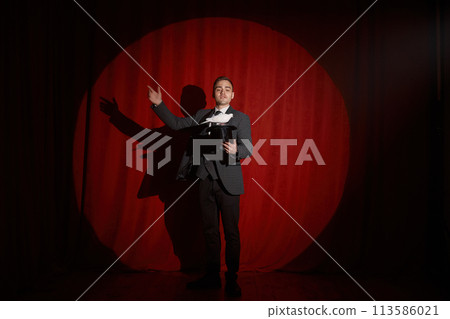 Magician with pigeon standing over red drapery in spotlight 113586021