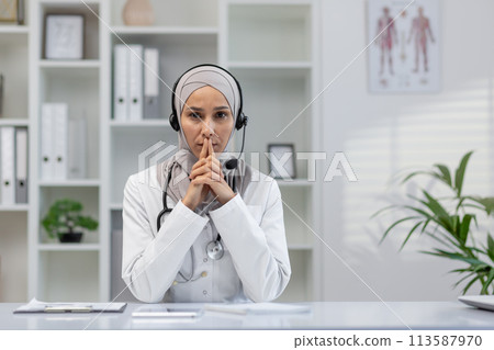 Focused female doctor wearing a hijab engaged in a serious video call, looking directly at the camera in a modern office setting. Focused female doctor wearing a hijab engaged in a serious video call, looking directly at the camera in a modern office setting. 113587970