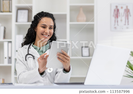 Cheerful female doctor in a white coat delights in using a digital tablet in her bright medical office, exemplifying professional happiness and digital healthcare. 113587996