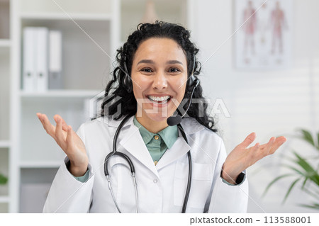 Cheerful female physician in a white coat using headphones for a video call. She looks into the camera with a friendly expression, offering medical advice and help. Cheerful female physician in a white coat using headphones for a video call. She looks into the camera with a friendly expression, offering medical advice and help. 113588001