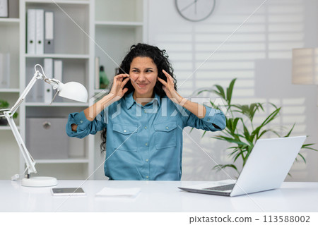 A professional woman in a blue shirt feeling overwhelmed by noise at her office desk with a laptop, plants, and a lamp 113588002