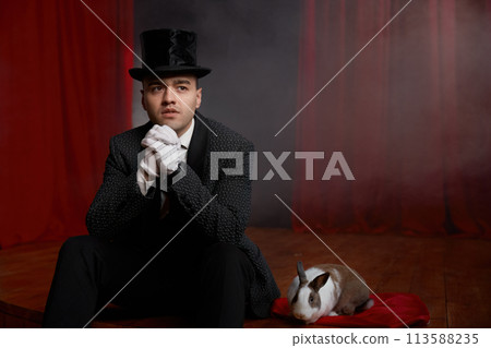 Pensive showman in suit sitting on theatre stage resting after magic show 113588235
