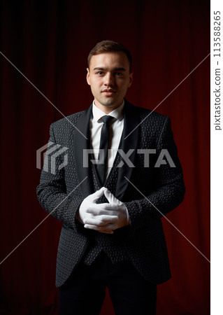 Magician wearing suit standing with cane on stage decorated red velvet curtain 113588265