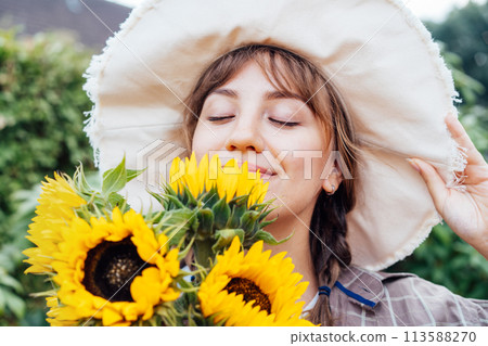 Close up Dreaming young female farmer woman with closed eyes holding and sniffing a sunflowers bouquet on the green garden background. Enjoy the moment. Rural, Cottage core lifestyle. 113588270