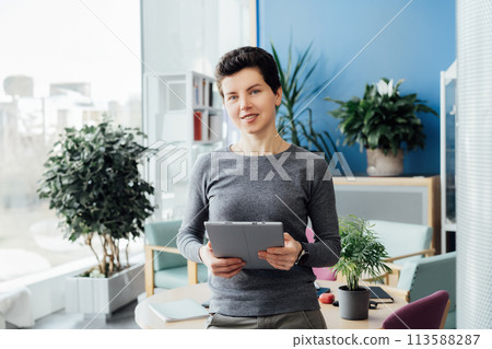 Portrait of smiling middle aged woman in casual outfit with digital tablet standing in company modern open space office, meeting room. Welcoming in office. Comfy workplace, selective focus. 113588287