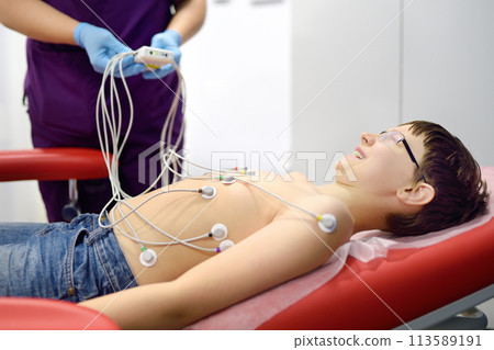 Child under ECG test with doctor. Boy patient lying in medical bed with vacuum sensors. A nurse performs an electrocardiogram of the heart in office of a medical clinic. 113589191
