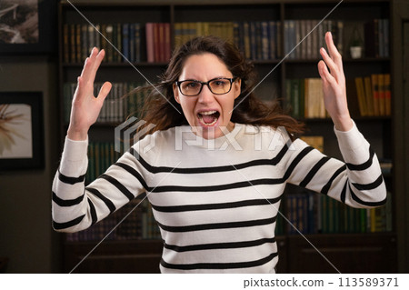 Portrait of an angry woman boss with glasses in the office looks at the camera and screams Portrait of an angry woman boss with glasses in the office looks at the camera and screams 113589371