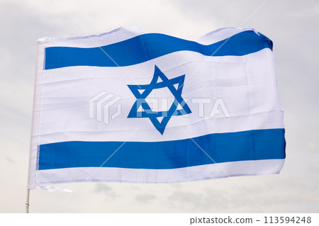 Canvas of Israeli flag flutters on flagpole against gray sky 113594248
