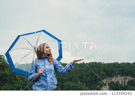 Young woman holding transparent blue umbrella outdoors in forest. Rainy weather day using umbrella. Woman with hand checking how long it will be raining  113594283
