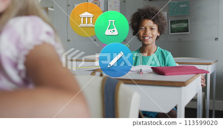 Image of school icons over smiling biracial schoolboy sitting at desk in class Image of school icons over smiling biracial schoolboy sitting at desk in class 113599020